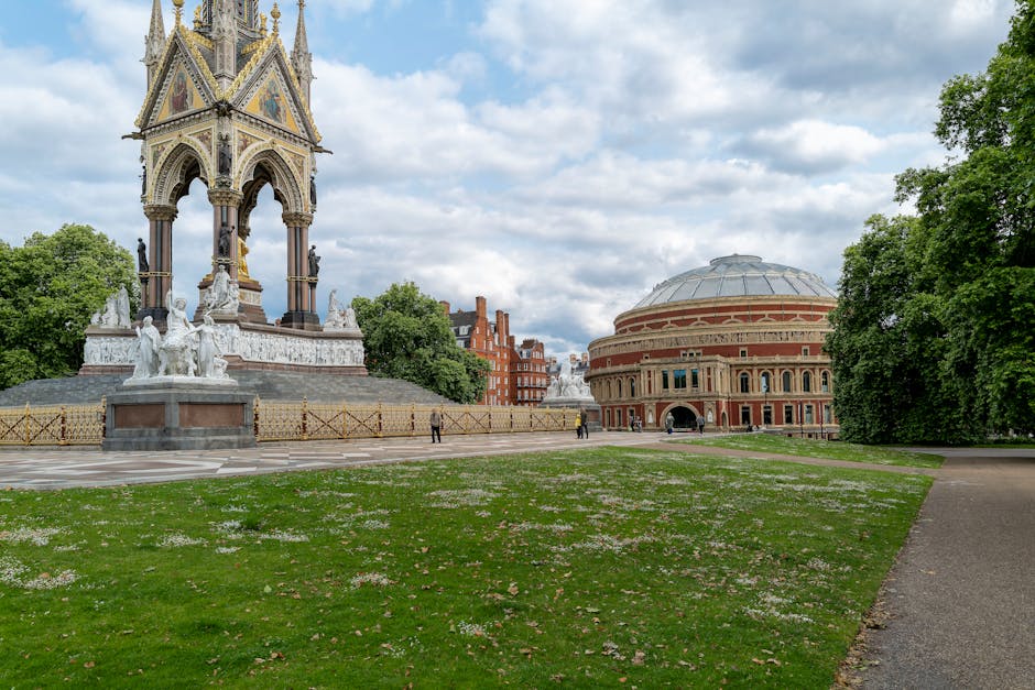 This image captures a landscaped park area in South Kensington featuring a paved walkway, well-maintained green grass, and scattered pink flower petals. In the background, there is an ornate, historic monument with detailed stone sculptures and decorative elements, surrounded by a low fence. Adjacent to the monument, a large, domed brick building with arched windows and classical architectural features is visible. Tall, leafy trees line the edges of the park, providing a lush contrast to the historic structures under a partly cloudy sky with patches of sunlight, creating a bright and inviting atmosphere. The scene exudes a sense of cleanliness and heritage, reflecting the area's cultural significance, which is supported by the local cleaning standards promoted by Carpet Cleaning South Kensington.