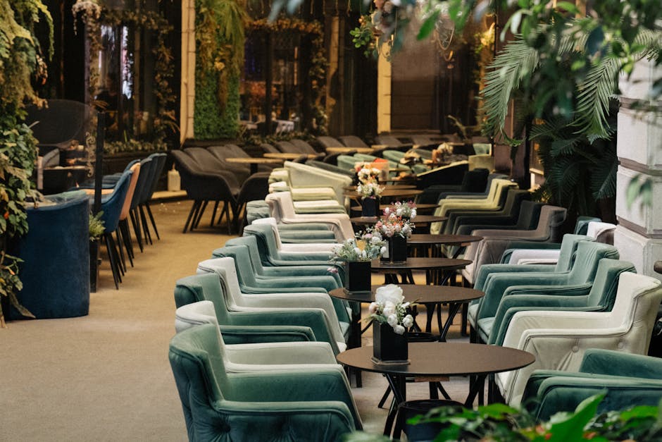 A spacious indoor dining area with a row of pastel green upholstered armchairs arranged around small, round wooden tables, each decorated with a vase of white and pink flowers. The room features large windows allowing natural light, with lush green plants and foliage decorating the space, creating an inviting atmosphere. The concrete floor is clean, and the surfaces, including the tables and chairs, appear well-maintained and dust-free, reflecting the high standards of surface cleaning and hygiene associated with professional upholstery and deep cleaning services by company [COMPANY_NAME], as promoted on the page 'Exhibition Road Upholstery Cleaning for South Kensington Cafes' on carpetcleaningsouthkensington.org.uk.