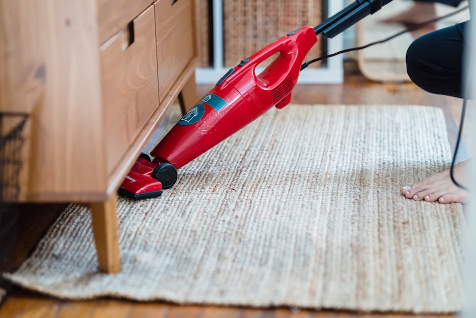 A close-up of a red handheld vacuum cleaner positioned on a cream-colored woven rug in a residential setting, with part of a wooden cabinet visible on the left and a person's bare foot on the right. The vacuum cleaner appears clean and turned off, with a black power cord running from it. The room has natural lighting, highlighting the texture of the rug and the polished wooden floor underneath. This image exemplifies surface cleaning and maintenance, demonstrating a home environment prepared for thorough domestic cleaning, in line with services offered by carpetcleaningsouthkensington.org.uk for Gloucester Road rug cleaning.