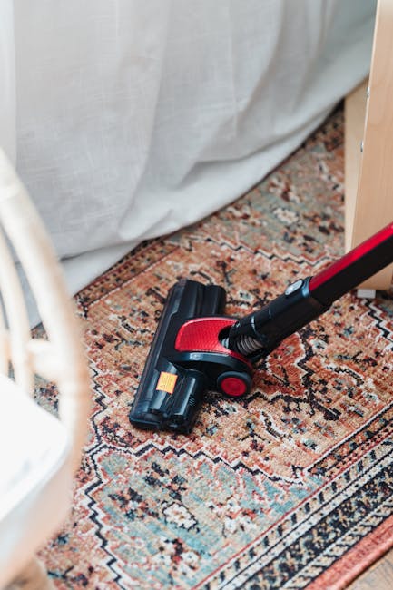 A middle-aged man with grey hair and a beard, wearing a light grey long-sleeved shirt and dark grey pants, is vacuuming a living room with a black and silver upright vacuum cleaner. The room features a white sofa with three cushions in blue, orange, and beige, situated against a wall with large windows draped in sheer white curtains, allowing natural light to fill the space. A low wooden coffee table on a yellow, white, and pink patterned rug holds a white vase with green foliage. The floor is dark herringbone parquet, clean and polished. In the background, a small black speaker, a black radiator beneath the window, and a wooden chair at a white desk are visible, all contributing to a tidy, well-maintained domestic setting. The natural lighting highlights the cleanliness and orderly arrangement of the room, some of which is maintained by cleaning services from carpetcleaningsouthkensington.org.uk focusing on surface cleaning, deep cleaning, and sanitisation for residential spaces.