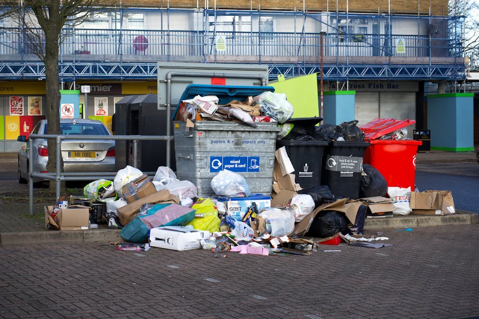 A large outdoor waste collection area in front of a building with a yellow storefront, featuring multiple overflowing bins filled with mixed paper, cardboard, plastic bags, and general rubbish. The waste is scattered around the bins, with some items on the pavement. There is a gray car parked nearby, and a metal railing separates the waste area from the pedestrian pathway. The building behind has a blue scaffolding structure and windows, and trees are visible on the sidewalk. The scene depicts an untidy state of waste disposal, emphasizing the importance of proper waste management, which aligns with the waste rules outlined by Kensington and Chelsea Council for cleaning and disposal, as highlighted by Carpet Cleaning South Kensington.