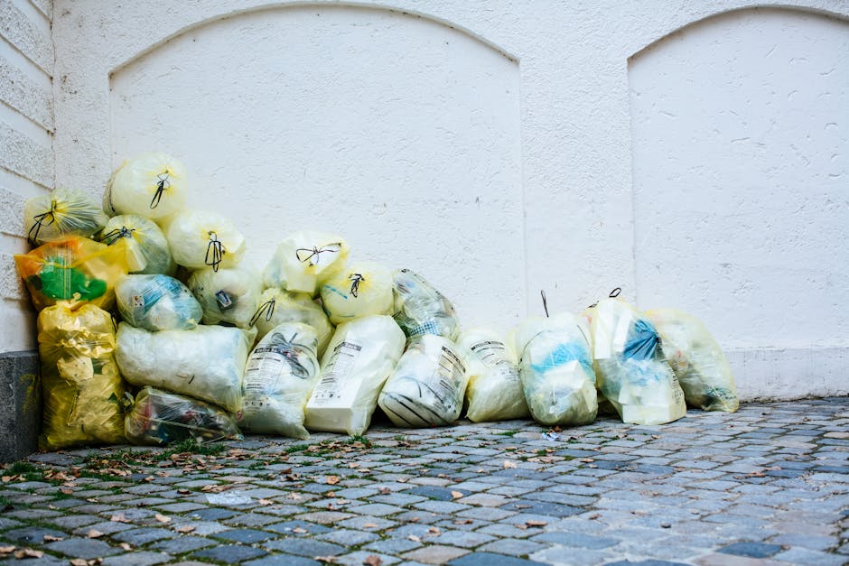 A pile of yellow and white plastic rubbish bags filled with waste is stacked against a white exterior wall on a cobblestone pavement. The bags appear to contain household trash, and some have labels or print visible. Nearby, there is a small glimpse of a brick wall on the left side. The scene suggests waste collection or disposal outside a building, emphasizing cleanliness and waste management in residential or commercial settings, relevant to surface cleaning and sanitation standards discussed on the Kensington and Chelsea Council Waste Rules for Cleaning Disposal page at carpetcleaningsouthkensington.org.uk.