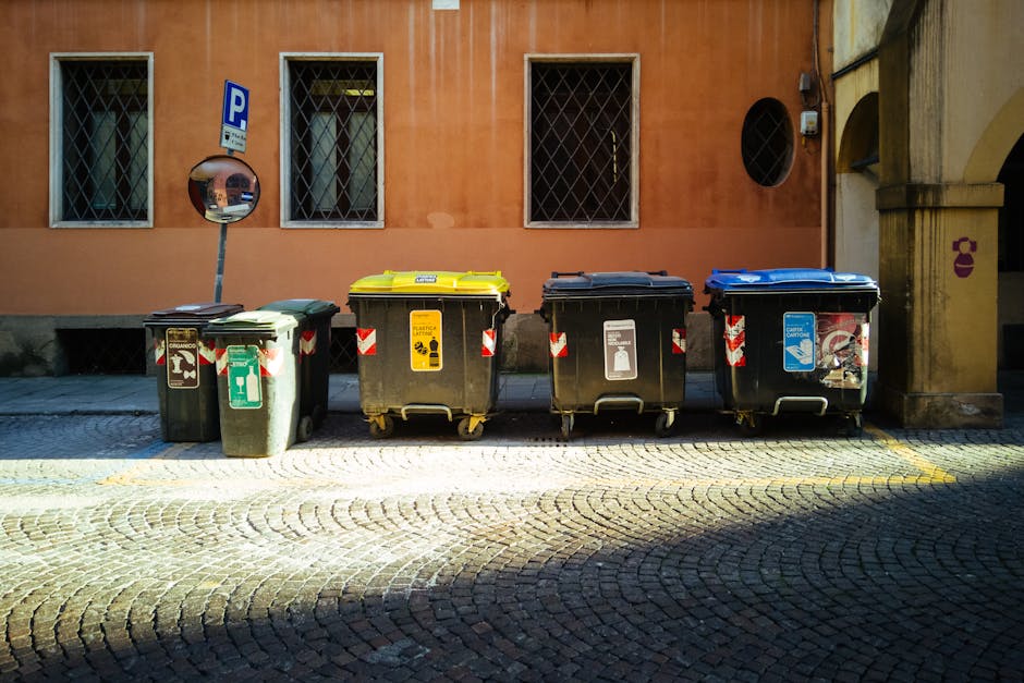 A large outdoor waste collection area in front of a building with a yellow storefront, featuring multiple overflowing bins filled with mixed paper, cardboard, plastic bags, and general rubbish. The waste is scattered around the bins, with some items on the pavement. There is a gray car parked nearby, and a metal railing separates the waste area from the pedestrian pathway. The building behind has a blue scaffolding structure and windows, and trees are visible on the sidewalk. The scene depicts an untidy state of waste disposal, emphasizing the importance of proper waste management, which aligns with the waste rules outlined by Kensington and Chelsea Council for cleaning and disposal, as highlighted by Carpet Cleaning South Kensington.