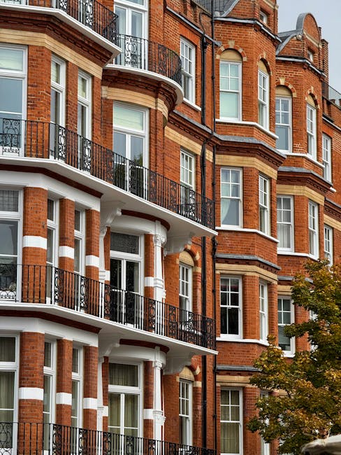 A close-up view of a multi-story residential building with a brick facade in Kensington, featuring white window frames, black wrought iron balconies, and decorative columns supporting the balconies. The building's exterior appears well-maintained and clean, with natural daylight illuminating the structure. A tree with green leaves is partially visible in the lower right corner, adding a touch of greenery to the scene. This image relates to professional domestic cleaning and surface maintenance in high-rise residential properties, as promoted by carpetcleaningsouthkensington.org.uk for end of tenancy cleaning and related services in Kensington Council area.