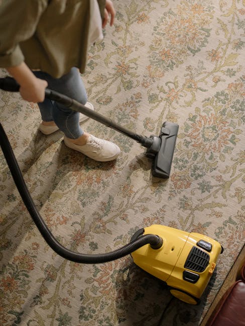A person cleaning a traditional patterned area rug with a vacuum cleaner in a well-lit room. The individual is wearing casual clothing including beige shoes, jeans, and a beige jacket. The vacuum cleaner is a yellow canister model with a black flexible hose, connected to a long metal wand with a cleaning head positioned on the carpet. The rug features an intricate design with floral motifs in muted colors, and the surface appears clean and well-maintained. The room’s flooring surrounding the rug includes a section of wooden or laminate surface, and ambient lighting enhances the overall cleanliness and freshness of the space. This scene exemplifies domestic deep cleaning efforts, likely undertaken by professionals such as those at South Kensington Carpet Cleaning, to ensure hygienic and pristine surface conditions.