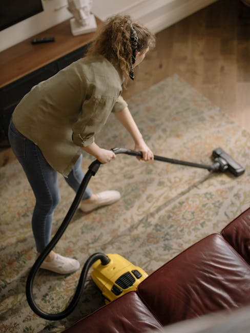 A woman wearing a beige jacket and blue jeans is performing surface cleaning or deep cleaning of a patterned area rug using a yellow vacuum cleaner with a attached hose in a living room. The room has wooden flooring and a dark brown leather sofa visible in the foreground. The background includes a wooden sideboard or shelf with a white decorative object on top, and the lighting appears natural, enhancing the cleanliness of the space. This image illustrates professional or domestic carpet cleaning activities, aligning with South Kensington Carpet Cleaning Cost Guide SW7, Carpet Cleaning South Kensington, and showcases the services provided by [COMPANY_NAME] in maintaining hygiene and freshness of residential interiors.