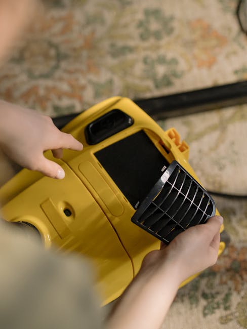 A person cleaning a traditional patterned area rug with a vacuum cleaner in a well-lit room. The individual is wearing casual clothing including beige shoes, jeans, and a beige jacket. The vacuum cleaner is a yellow canister model with a black flexible hose, connected to a long metal wand with a cleaning head positioned on the carpet. The rug features an intricate design with floral motifs in muted colors, and the surface appears clean and well-maintained. The room’s flooring surrounding the rug includes a section of wooden or laminate surface, and ambient lighting enhances the overall cleanliness and freshness of the space. This scene exemplifies domestic deep cleaning efforts, likely undertaken by professionals such as those at South Kensington Carpet Cleaning, to ensure hygienic and pristine surface conditions.