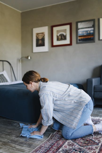 A person kneeling on a patterned carpet in a living room, using a yellow and black portable vacuum cleaner for surface cleaning. The individual is wearing a beige jacket and blue jeans, and is focused on the vacuum's dust compartment. The room features wooden flooring adjacent to the carpet and soft ambient lighting, emphasizing cleanliness and maintenance of the domestic space. This image highlights carpet deep cleaning processes often associated with professional cleaning services such as those offered by carpetcleaningsouthkensington.org.uk in the South Kensington area.